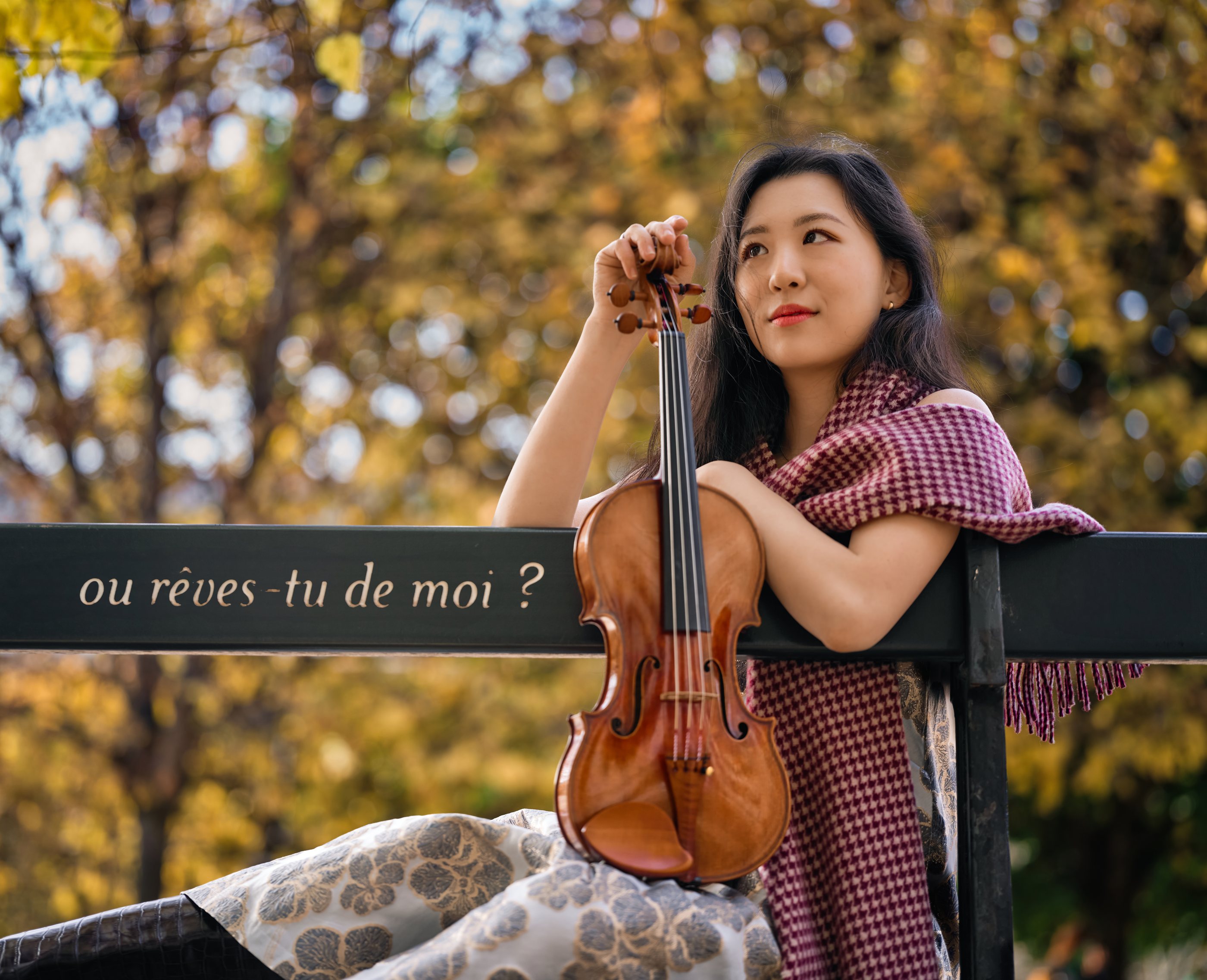 Anna Lee seated on a park bench with her violin among autumn leaves