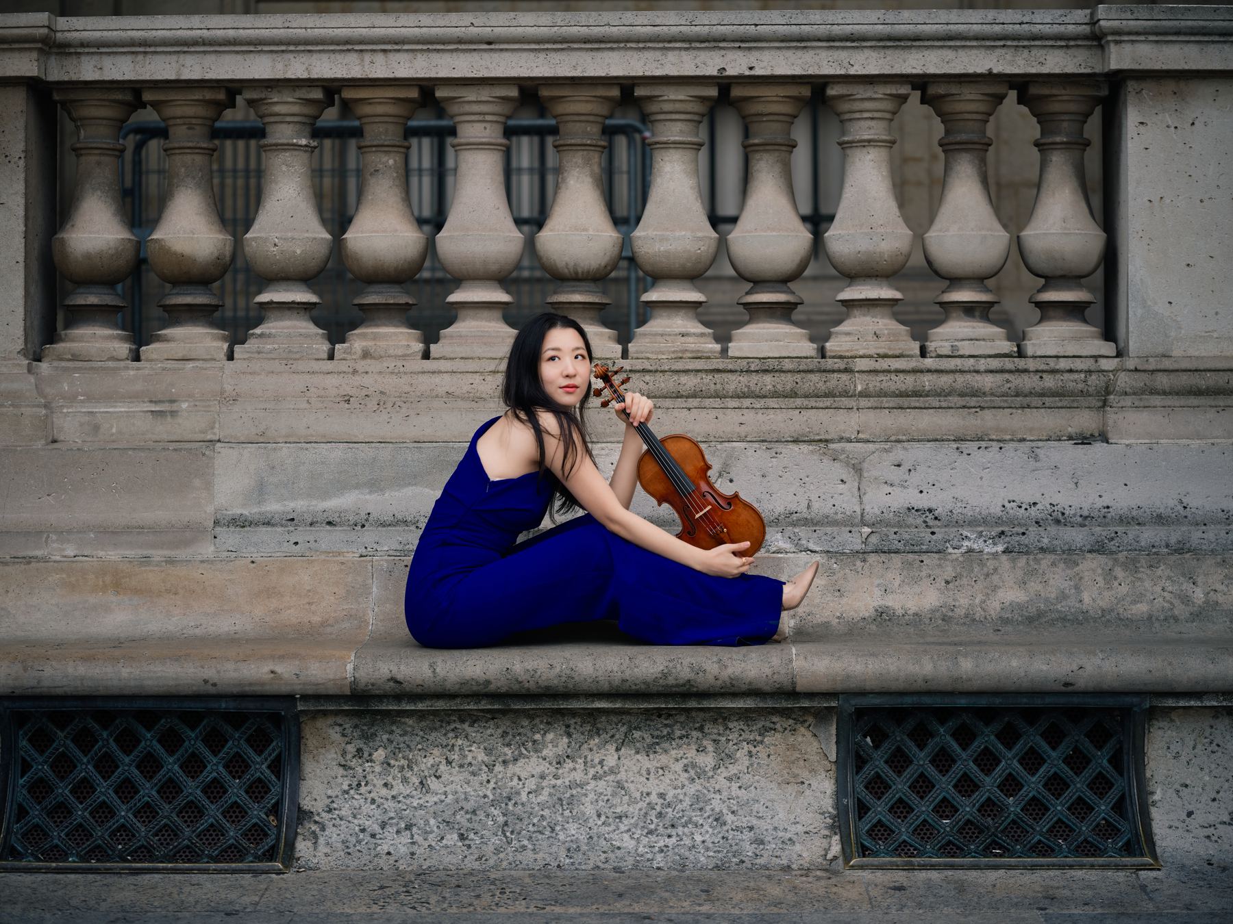 Anna Lee in blue seated with her violin against classical stonework